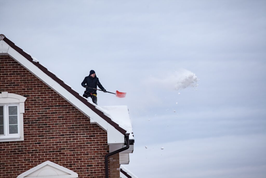 A man on a house rooftop carefully removing snow using a shovel